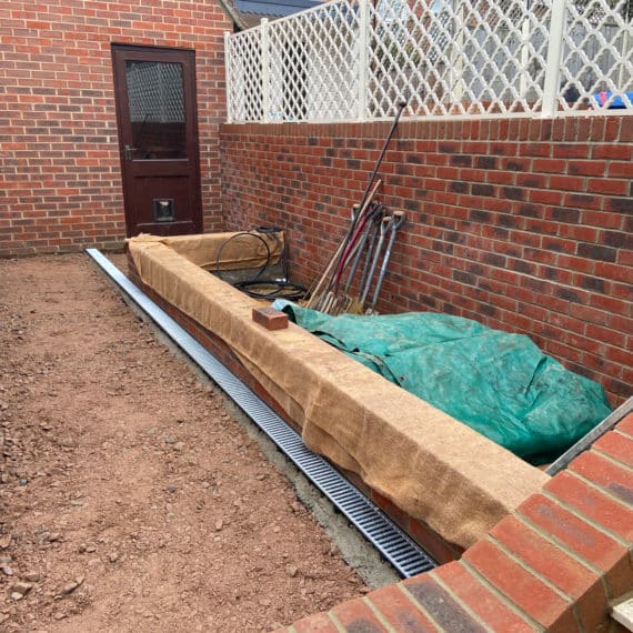 partially landscaped patio area with tall retaining wall to the right, brick built raised bed adjoining the wall is partially constructed